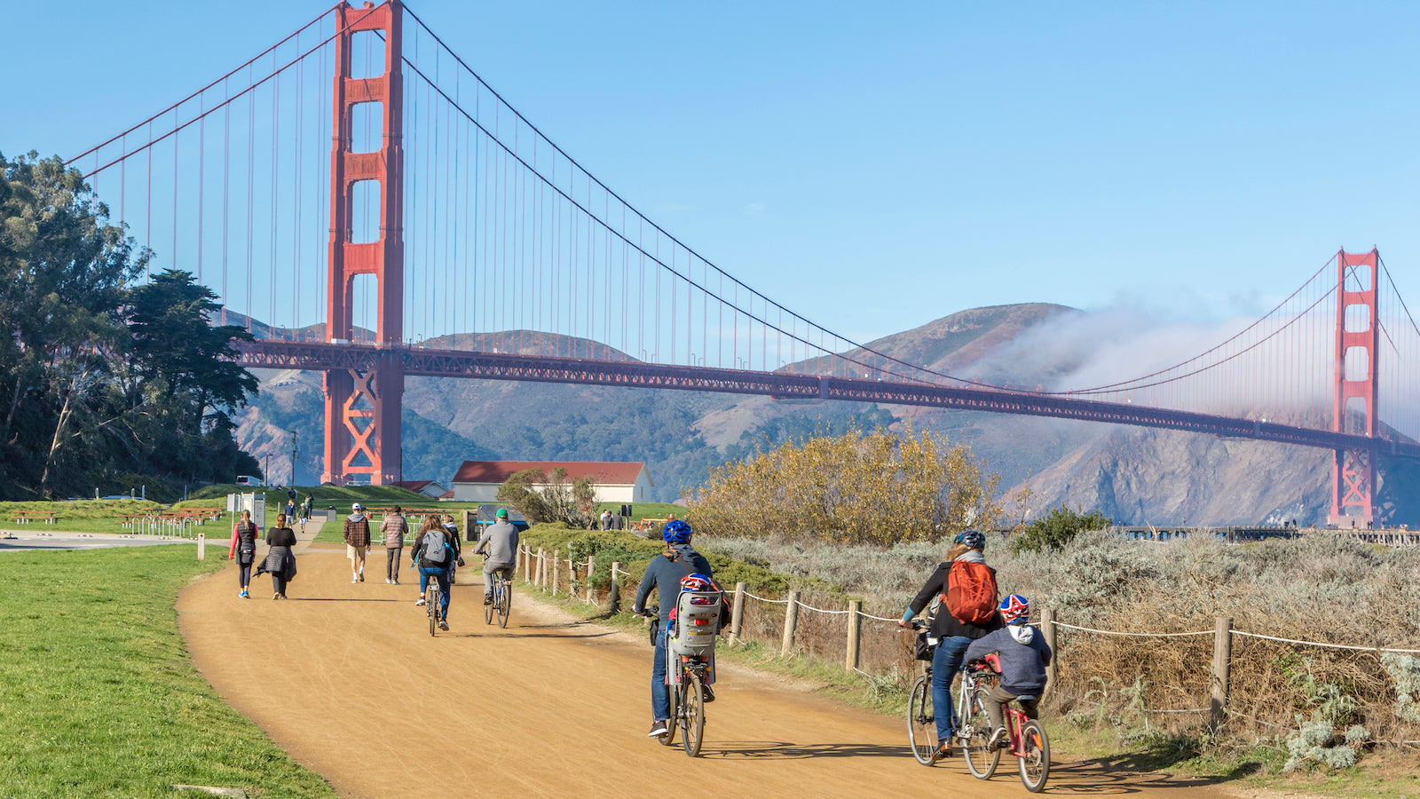 A view towards the Warming Hut Bookstore and Cafe on a sunny day. Bicyclists and walkers are busy in the foreground, while the Golden Gate Bridge rises above the misty Marin Headlands in the background.
