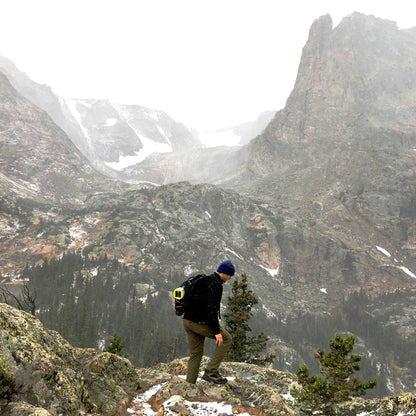 A man hikes in high altitude scenic mountains covered in snow. He wears a beanie and carries a backpack with a rechargeable Luci solar light attached.